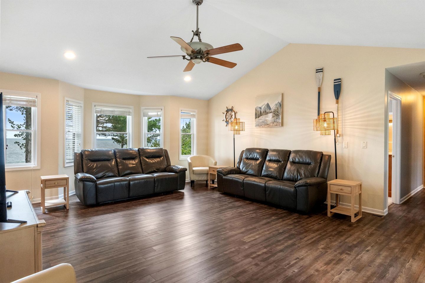 A spacious living room with a dark brown leather sofa, a white armchair, a wooden coffee table, and a ceiling fan.