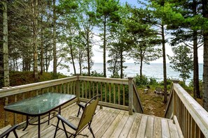 Terraza de madera con una mesa y sillas de vidrio, con vista a un bosque y al océano.