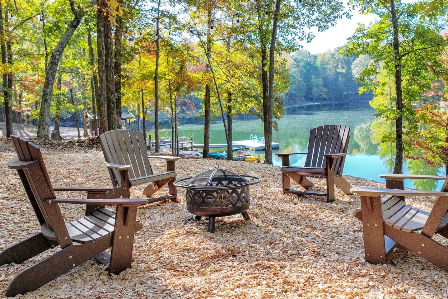 A lakeside seating area with Adirondack chairs and a fire pit, surrounded by trees and a view of the lake.