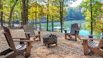 A lakeside seating area with Adirondack chairs and a fire pit, surrounded by trees and a view of the lake.