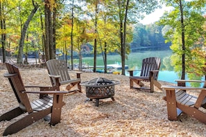 A lakeside seating area with Adirondack chairs and a fire pit, surrounded by trees and a view of the lake.
