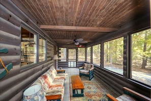 Ein Blockhauskamin mit einer Couch, Sesseln, einem Tisch und großen Fenstern mit Blick auf den Wald.