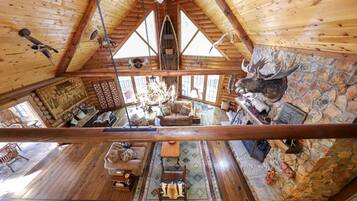 A log cabin interior with a high ceiling, wooden walls, a stone fireplace, and a mounted deer head.