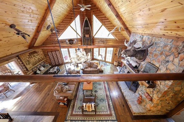 A log cabin interior with a high ceiling, wooden walls, a stone fireplace, and a mounted deer head.