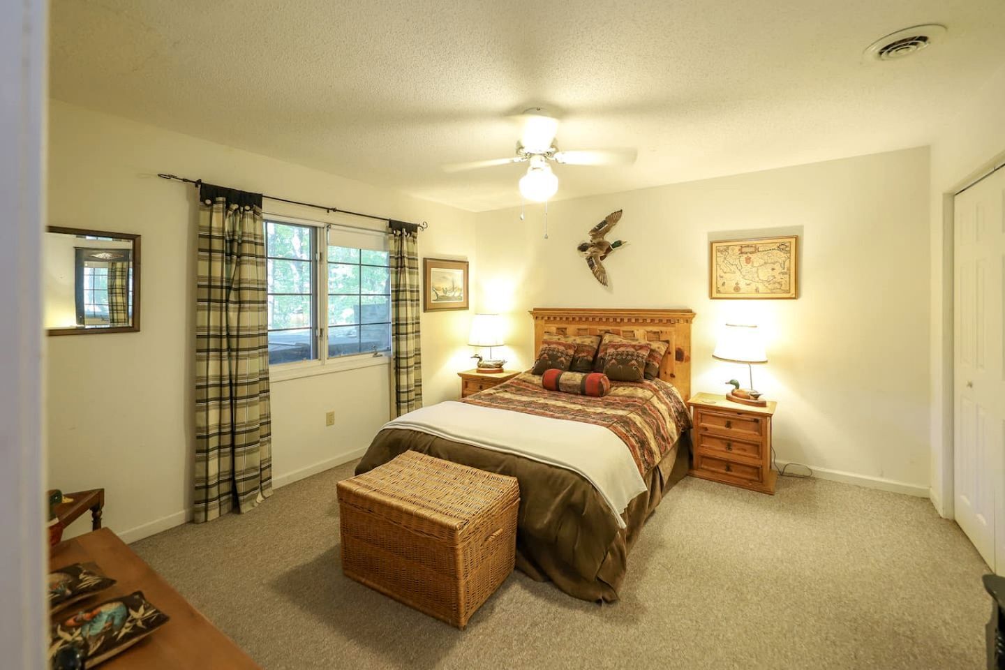 A bedroom with a bed, two bedside tables, a nightstand, a window with curtains, a ceiling fan, and a wall-mounted bird decoration.
