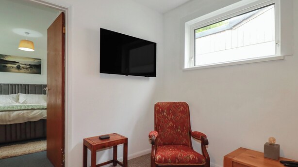 Ein Zimmer mit einem Bett, einem an der Wand befestigten Fernseher, einem Fenster, einem Sessel, einem kleinen Tisch und einer Tür zu einem anderen Raum.
