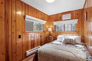 Ein Schlafzimmer mit Holzwänden, einem Bett, zwei Fenstern mit weißen Jalousien, einem Nachttisch und einem Wandheizkörper.