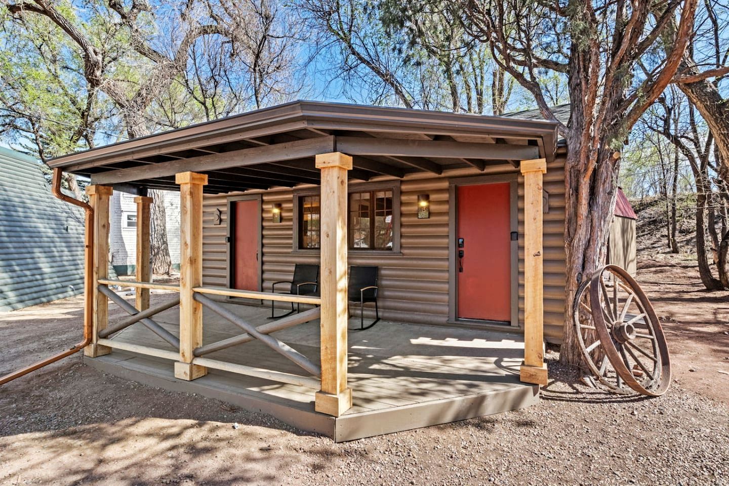 A rustic cabin with a covered porch, a red door, and a large wheel.