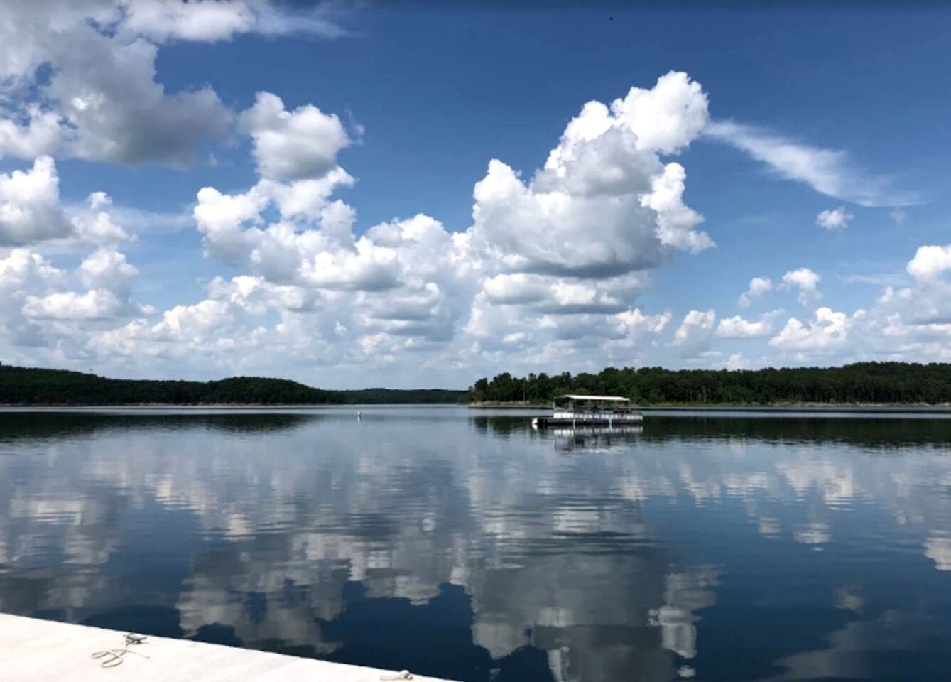A calm lake with a dock and a boat, surrounded by trees and a clear sky.