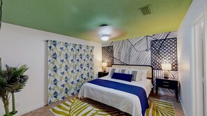Ein Schlafzimmer mit einem Bett, einem Nachttisch, einer Lampe, einem gemusterten Vorhang und einer Pflanze.