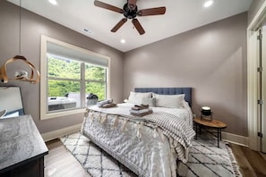 A bedroom with a bed, a ceiling fan, a window with a view, a bedside table, and a rug. - Mountain Hideaway with Waterfall Views & Relaxing Hammocks in Chimney Rock, North Carolina (Chimney Rock)