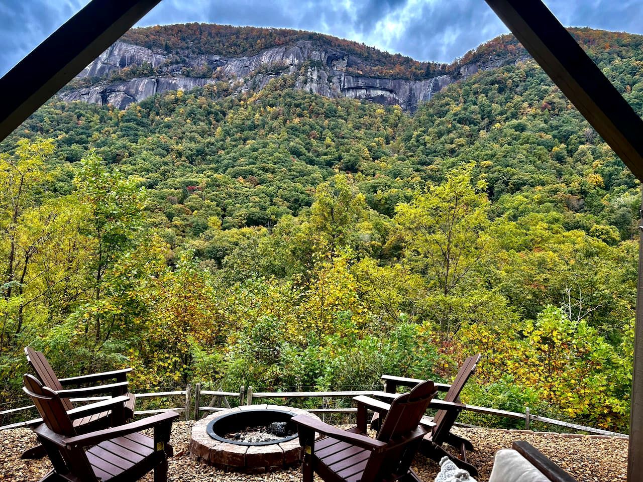 A view from a covered area with a fire pit and wooden chairs, overlooking a forested mountain.