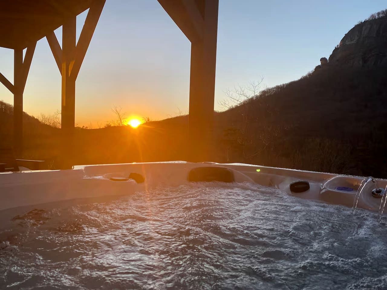 Un jacuzzi bajo un puente con agua, un atardecer al fondo y colinas.