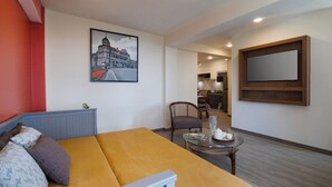 Moderni hotellihuone, jossa on vuodesohva, ruokapöytä, tuoli, keittiöalue ja suuri seinälle asennettu televisio.