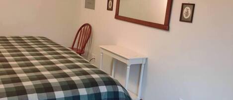 A bedroom with a bed, a chair, a desk, a mirror, and wall-mounted hangers.