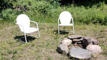 Dos sillas blancas y un hogar de piedra en un área cubierta de césped, con árboles al fondo.