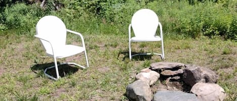 Deux chaises blanches et un foyer en pierre, le tout installé dans un espace verdoyant, avec des arbres en arrière-plan.