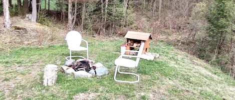 A fire pit with logs, a white chair, and a birdhouse in a grassy outdoor area.