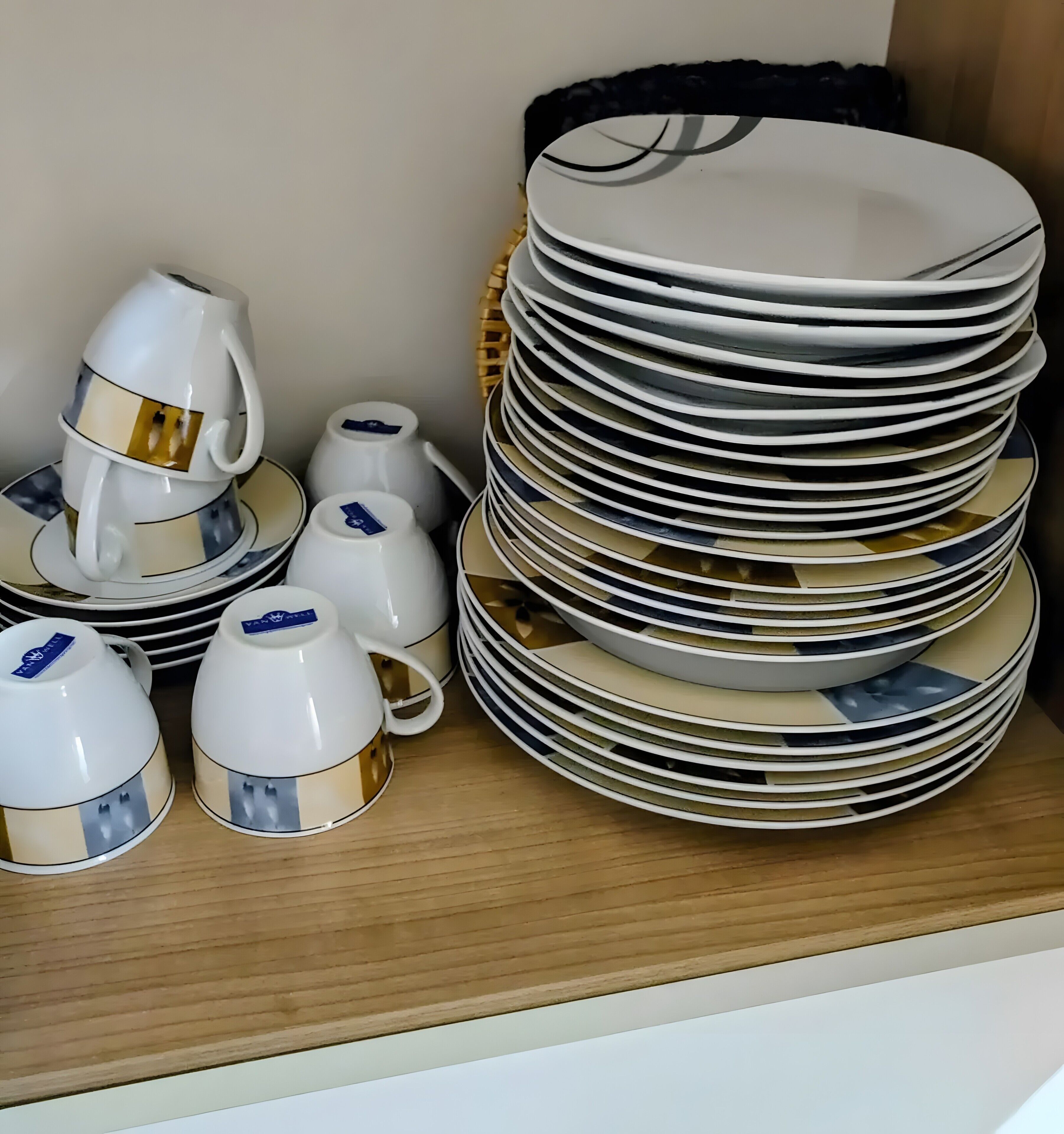 Une pile d’assiettes blanches ornées de motifs dorés, une théière blanche et des tasses assorties, le tout posé sur une étagère en bois.