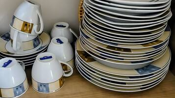 Une pile d’assiettes blanches ornées de motifs dorés, une théière blanche et des tasses assorties, le tout posé sur une étagère en bois.