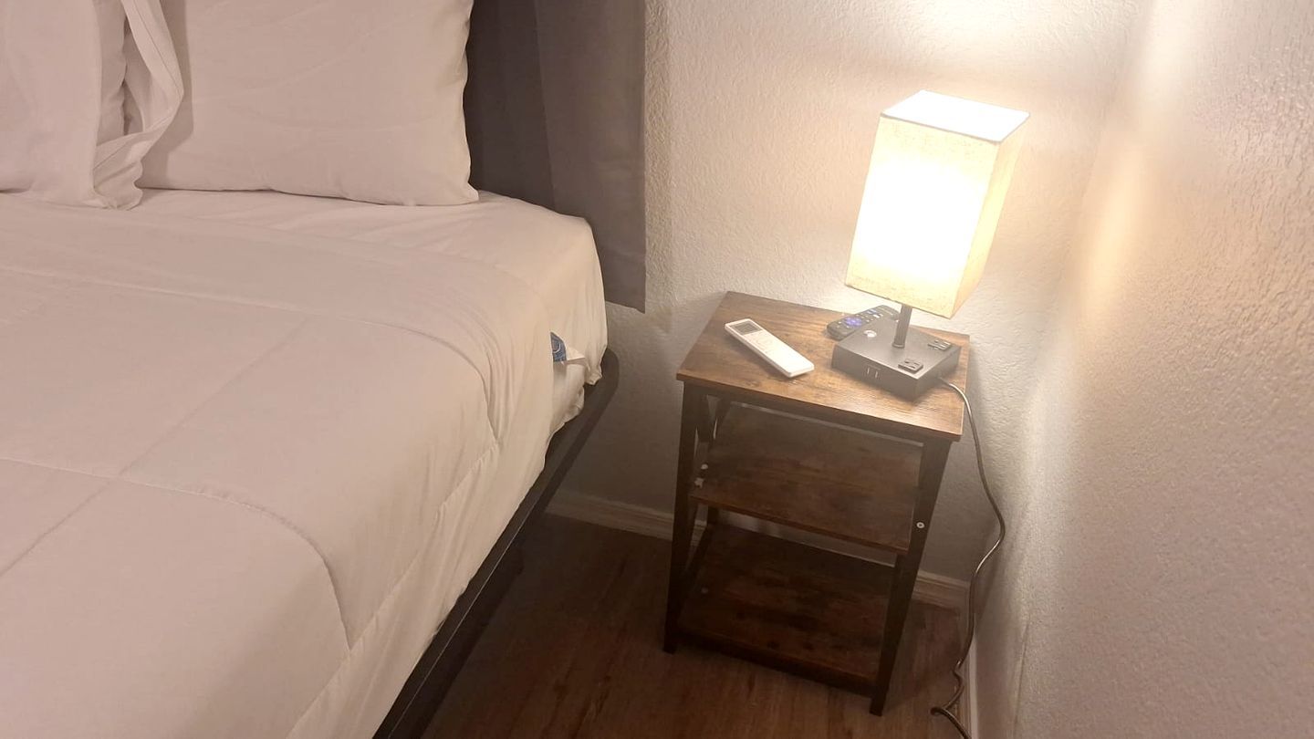 A hotel room with a bed, a bedside table with a lamp, and a power outlet.
