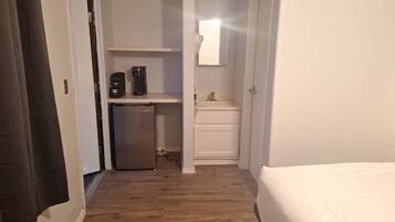 A hotel room with a bed, a small kitchenette with a refrigerator and coffee maker, a bathroom with a mirror, and a wooden floor.
