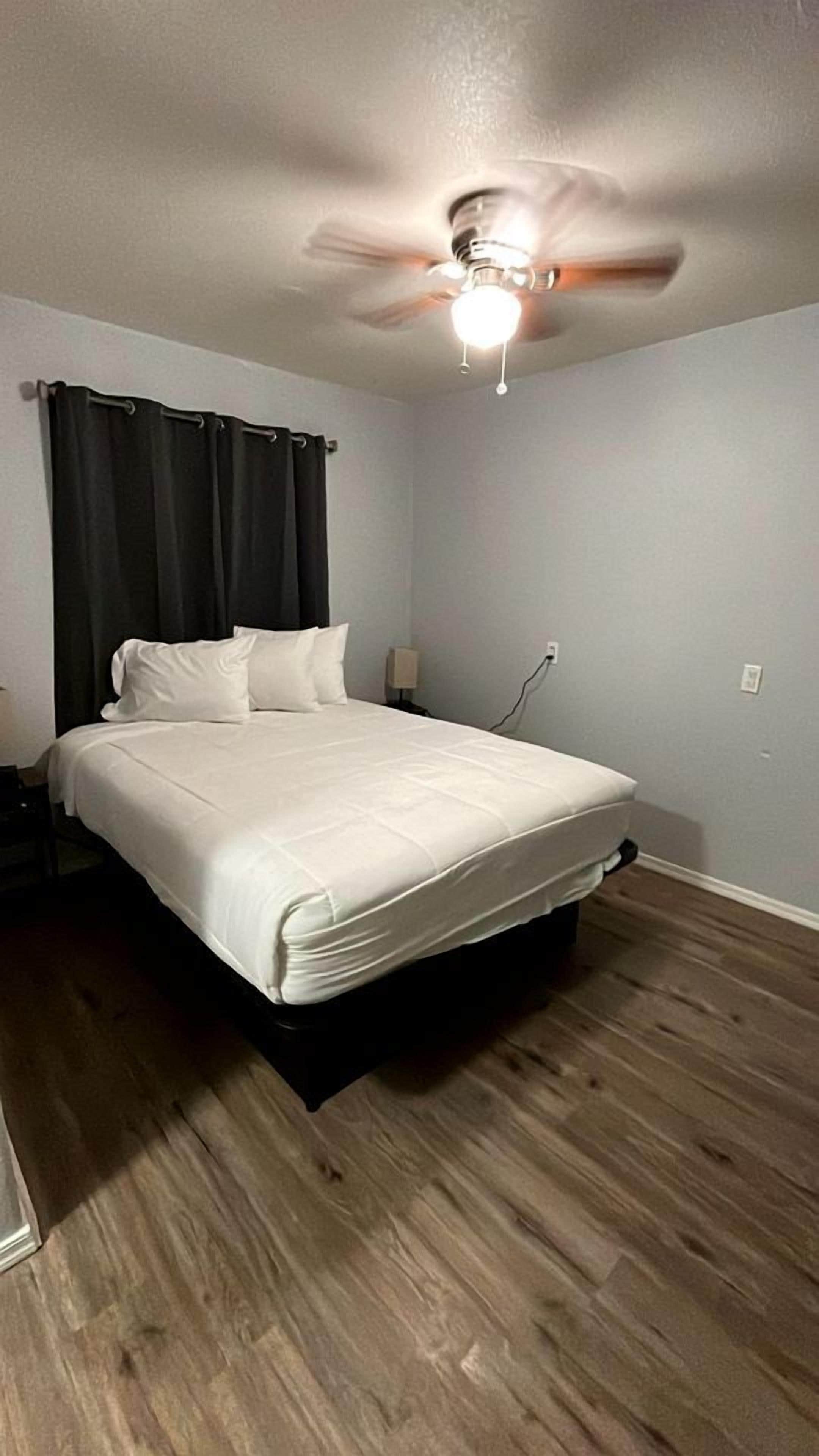 A hotel room with a bed, a ceiling fan, a nightstand, and a wall socket.