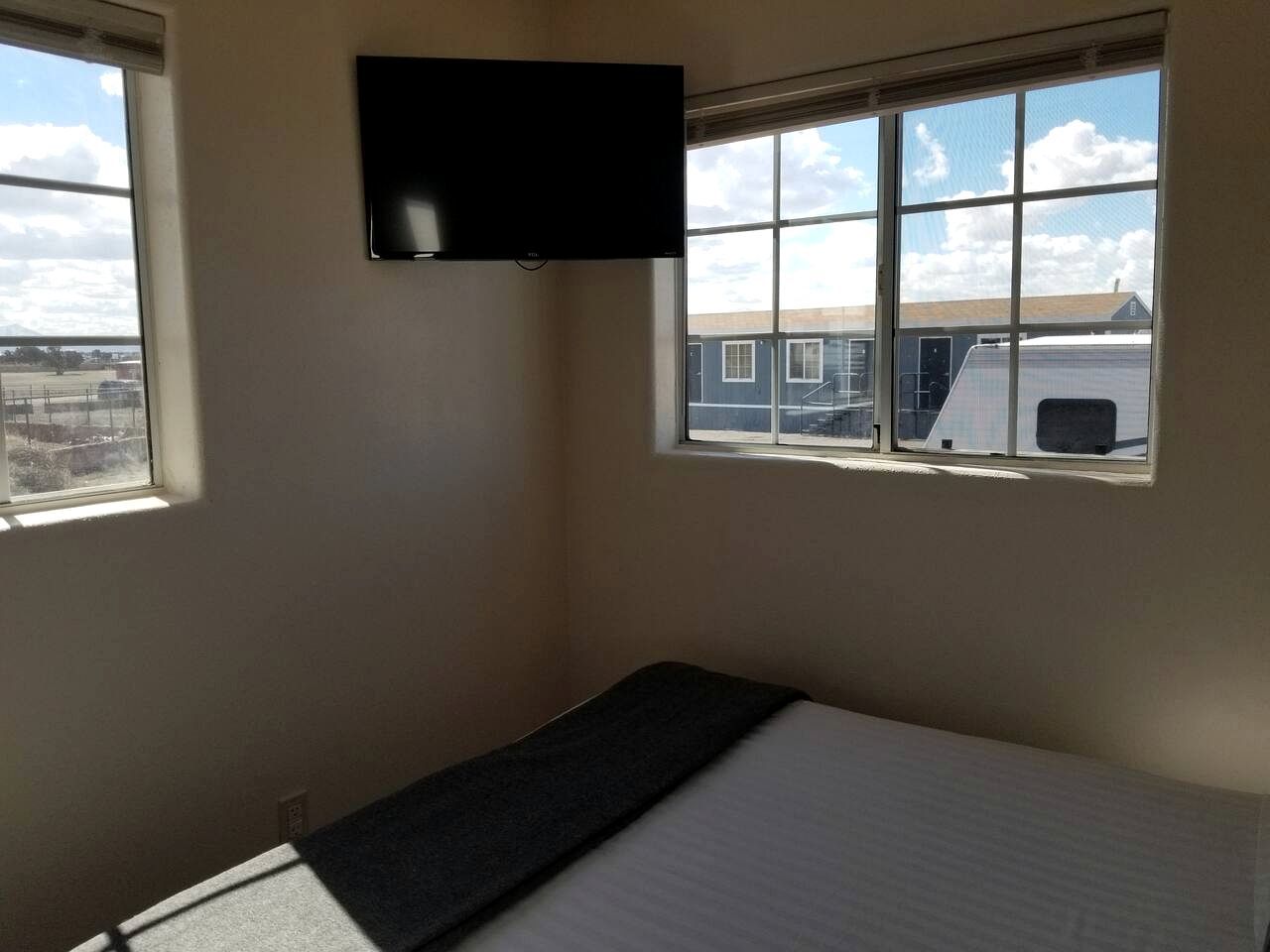 Una habitación pequeña con una cama, un televisor montado en la pared y una ventana con vistas a edificios.