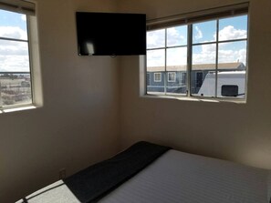 Una habitación pequeña con una cama, un televisor montado en la pared y una ventana con vistas a edificios.