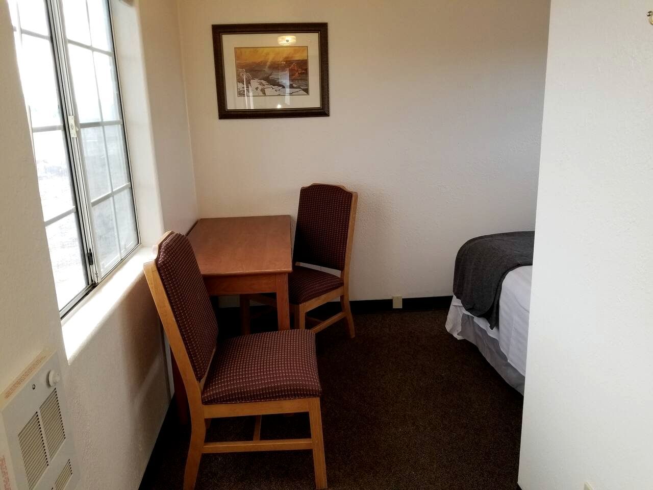 A small room with a table and two chairs, a bed, a window, and a framed picture on the wall.