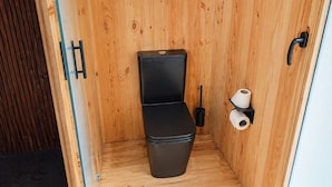 A compact wooden room with a black toilet, a toilet brush, and two rolls of toilet paper.