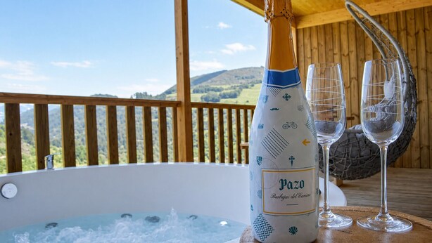 A bottle of Pazo Barbeitas del Camino champagne, two glasses, and a hot tub on a wooden deck with a view of hills.