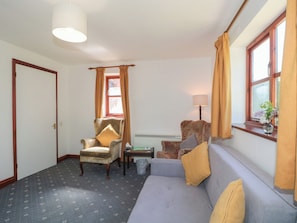 A room with a grey sofa, a patterned armchair, a small table, a lamp, and a window with curtains. - Walk Close (Gloucester)