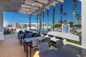 A covered outdoor area with tables and chairs, a pool, and palm trees.