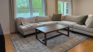 Een woonkamer met een hoekbank, een salontafel en grote ramen met gordijnen.