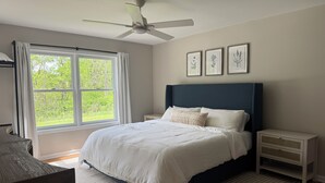 Een slaapkamer met een bed, nachtkastje, raambord, plafondventilator, raam met gordijnen en ingelijste botanische prints aan de muur.