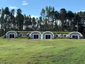 Una hilera de tres edificios modernos, semicirculares, con puertas grandes, ubicada en un campo cubierto de hierba y con un bosque al fondo.