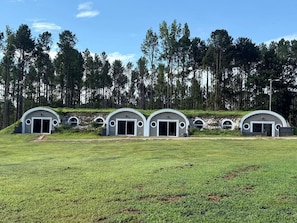 A row of three modern, semi-circular buildings with large doors, set in a grassy field with a forest in the background.