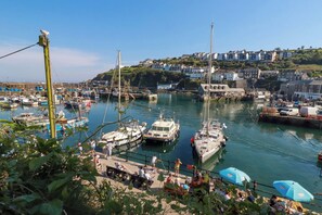 A marina with boats docked, a dock with people, and houses on a hill in the background. - Trewollock (St Austell)