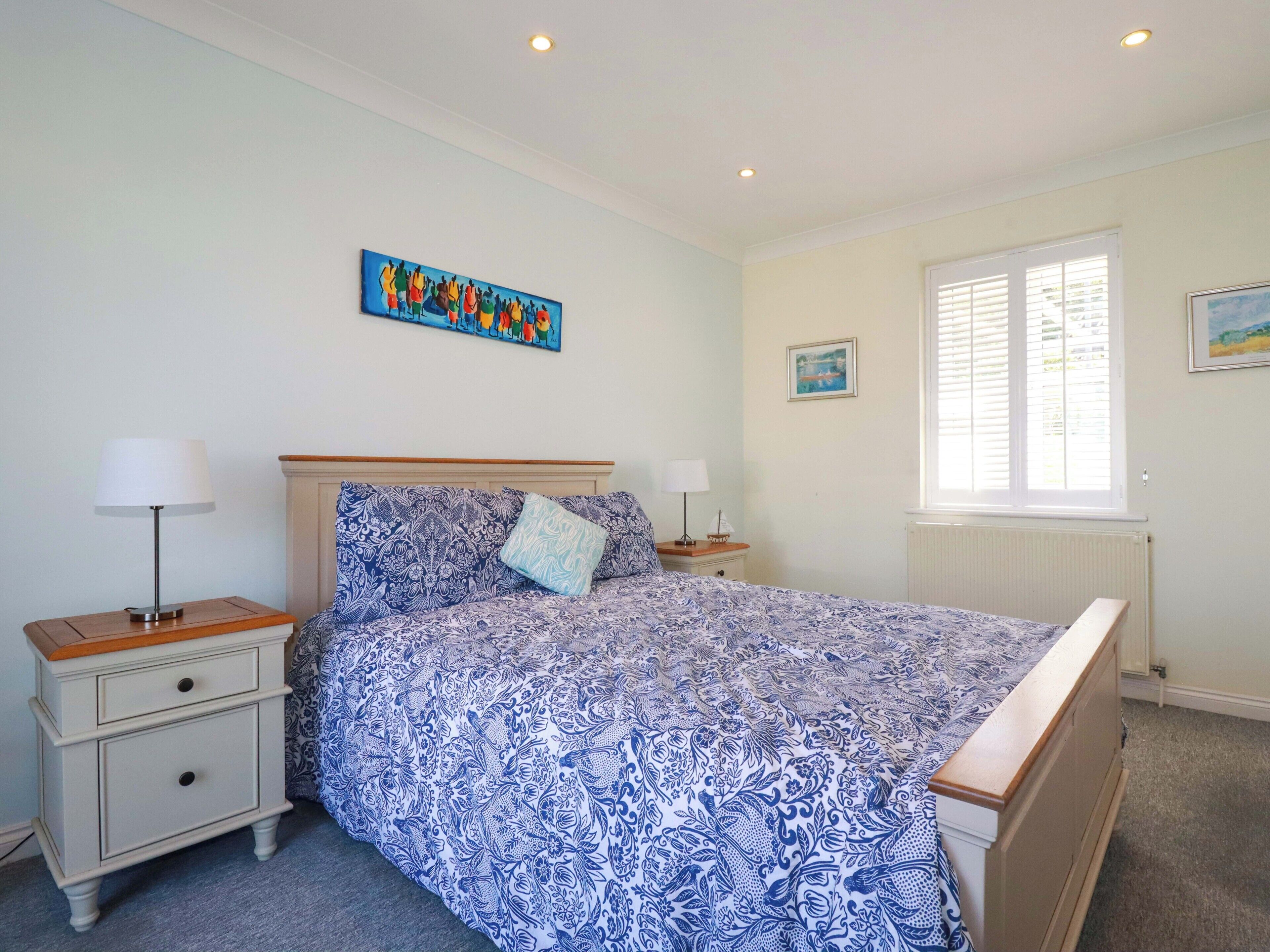 A bedroom with a bed, two bedside tables, a window with blinds, and a painting on the wall.