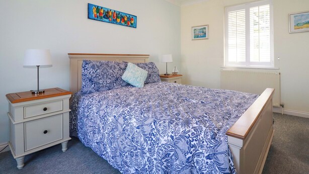 A bedroom with a bed, two bedside tables, a window with blinds, and a painting on the wall. - Trewollock (St Austell)