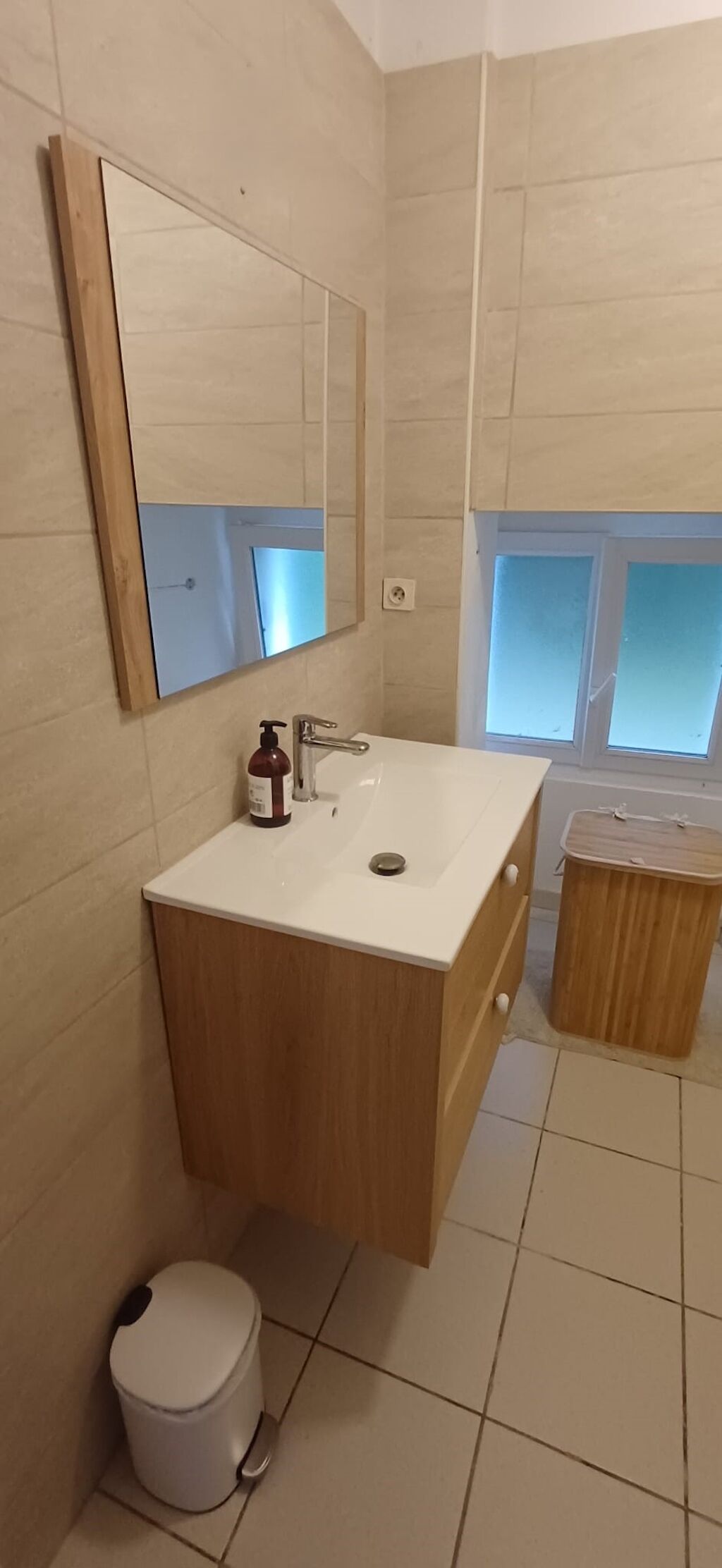 Une salle de bain avec un lavabo blanc, un meuble-lavabo en bois, un miroir, une fenêtre et une poubelle.