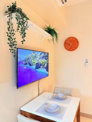 Ein Essbereich mit Fernseher, der ein Landschaftsbild zeigt, einer Uhr und einer Pflanze an der Wand.
