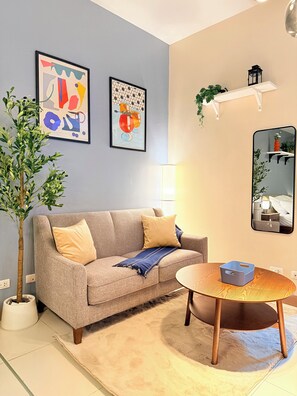 Ein Wohnzimmer mit einer Couch, einem runden Holztisch, einem Blumentopf und zwei gerahmten Kunstwerken an der Wand.