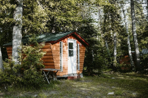 Una cabaña de troncos con un porche pequeño y una mesa, rodeada de un bosque denso.