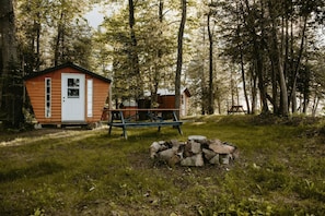 Une cabane rustique avec une façade en bois, un petit porche et une table de pique-nique, située dans une zone boisée.