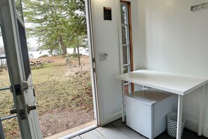 A room with a white desk, a small refrigerator, and a door leading to an outdoor area with trees and a picnic table.