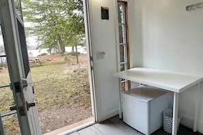 Une piĂšce avec un bureau blanc, un petit rĂ©frigĂ©rateur et une porte donnant sur un espace extĂ©rieur Ă©quipĂ© dâune table de pique-nique et bordĂ© dâarbres.