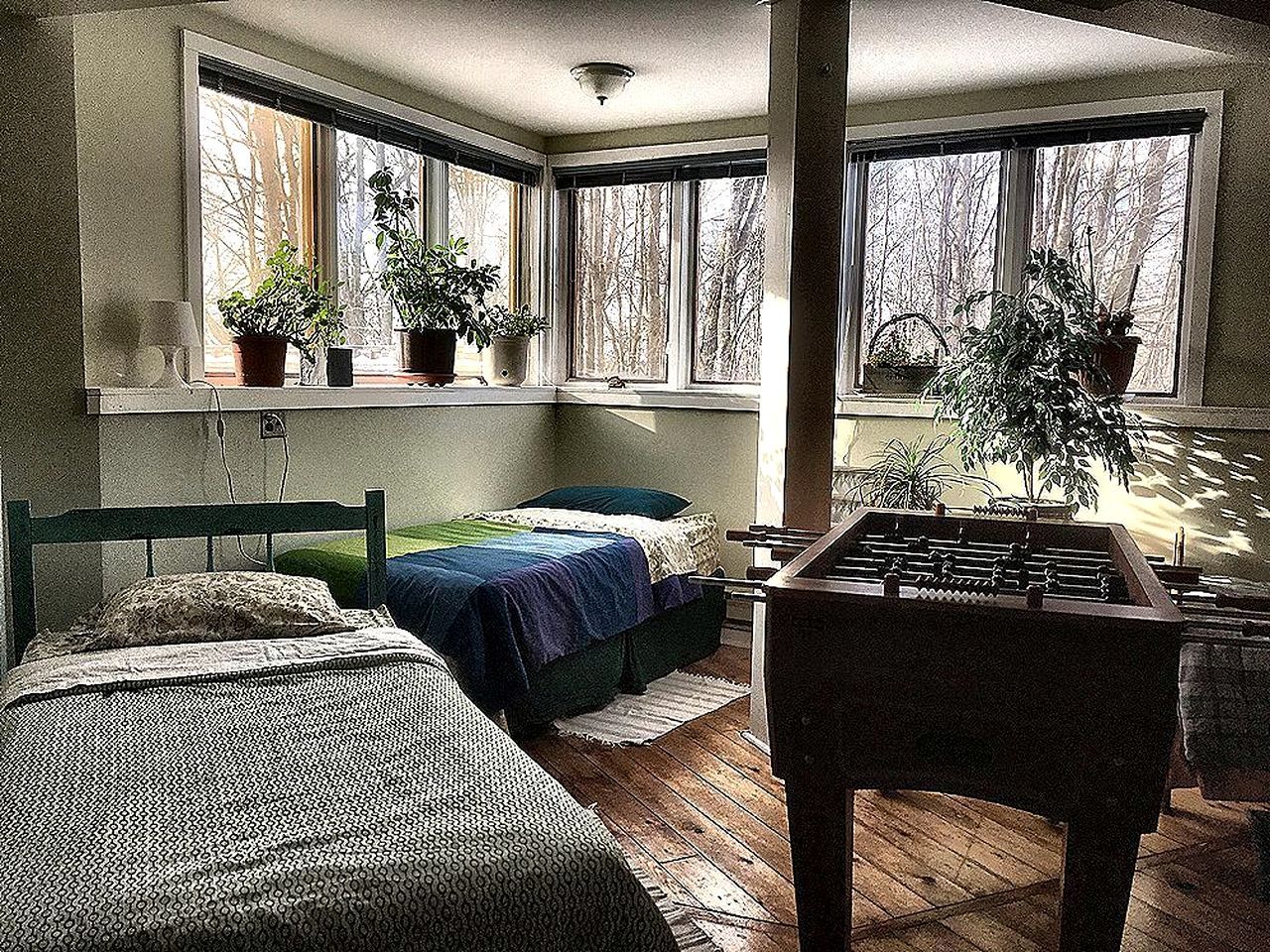 Una habitación con dos camas, una mesa de fútbol y ventanas amplias con plantas en macetas.