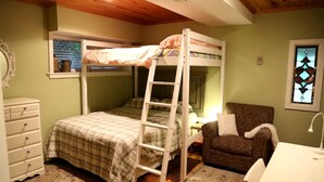 Un lit superposé avec une échelle, une chaise, un bureau et une commode dans une pièce dotée de poutres apparentes en bois au plafond et d’une fenêtre avec des stores.